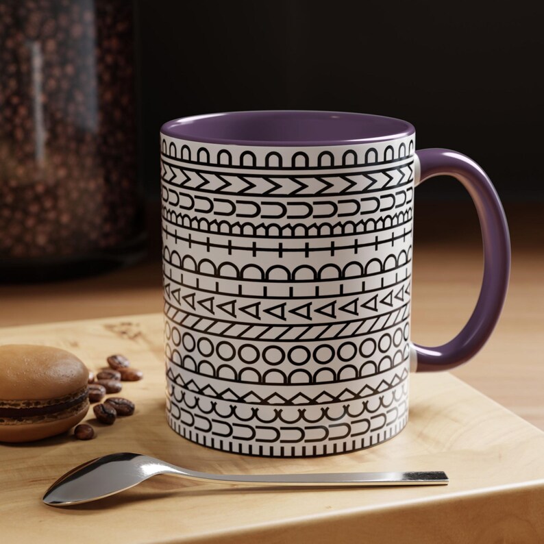 May include: A ceramic mug with a purple interior and handle, featuring a white exterior decorated with horizontal bands of black geometric patterns including circles, triangles, lines, and arches, arranged in a repeating, stacked design.