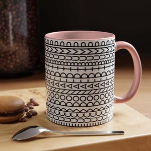 May include: A ceramic mug with a light pink interior and handle, featuring a white exterior decorated with horizontal bands of black geometric patterns, including lines, triangles, circles, and semicircles arranged in a repeating design.