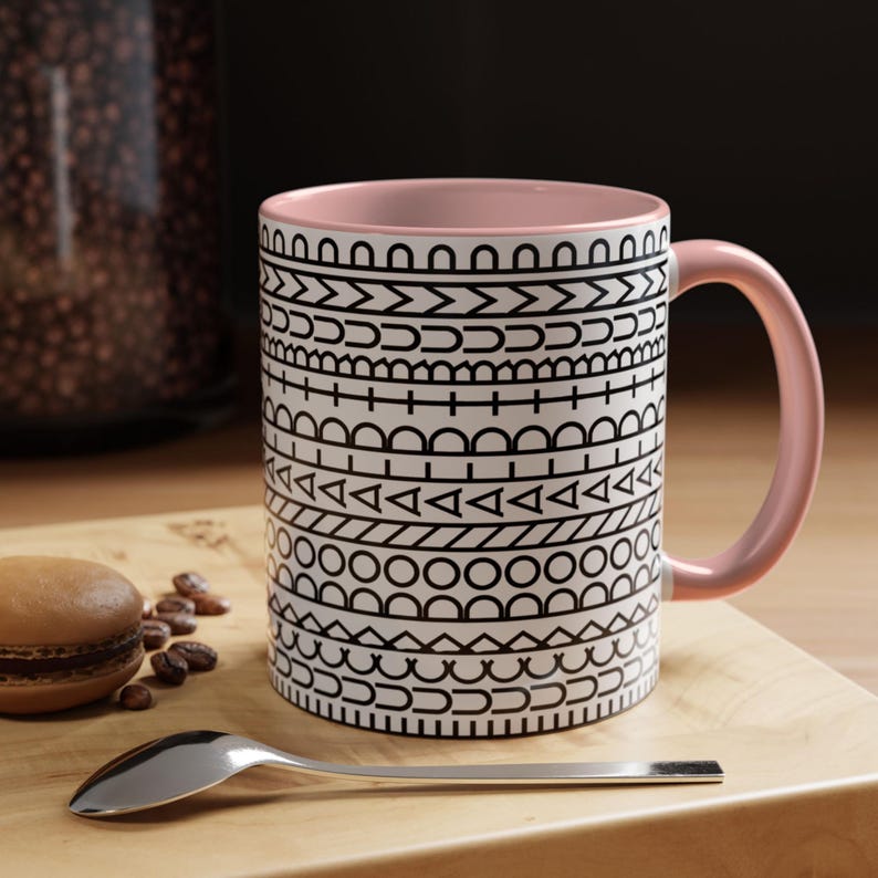 May include: A ceramic mug with a light pink interior and handle, featuring a white exterior decorated with horizontal bands of black geometric patterns, including lines, triangles, circles, and semicircles arranged in a repeating design.