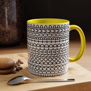 May include: A ceramic mug with a bright yellow interior and handle, featuring a white exterior decorated with horizontal bands of black geometric patterns including circles, triangles, lines, and arches, arranged in a repeating linear design.