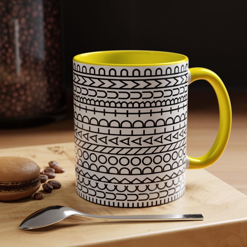 May include: A ceramic mug with a bright yellow interior and handle, featuring a white exterior decorated with horizontal bands of black geometric patterns including circles, triangles, lines, and arches, arranged in a repeating linear design.