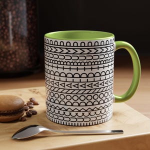 May include: A ceramic mug with a bright lime green interior and handle. The exterior features a repeating black and white geometric pattern consisting of horizontal bands of semicircles, triangles, circles, and lines arranged in a stacked design.