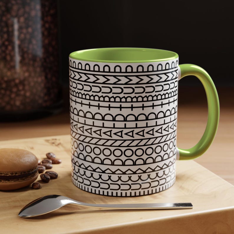 May include: A ceramic mug with a bright lime green interior and handle. The exterior features a repeating black and white geometric pattern consisting of horizontal bands of semicircles, triangles, circles, and lines arranged in a stacked design.