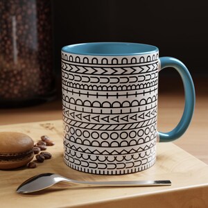 May include: A ceramic mug with a light blue interior and handle, featuring a white exterior decorated with horizontal bands of black geometric patterns including triangles, circles, and lines. The mug is positioned next to a macaron and a silver spoon.
