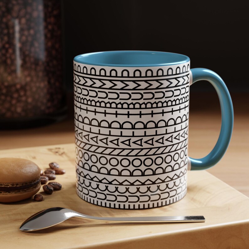 May include: A ceramic mug with a light blue interior and handle, featuring a white exterior decorated with horizontal bands of black geometric patterns including triangles, circles, and lines. The mug is positioned next to a macaron and a silver spoon.