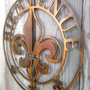 May include: A round metal sign featuring a fleur-de-lis design in the centre and the word "Bienvenue" around the edge. The sign is painted in a gold colour with a distressed finish.