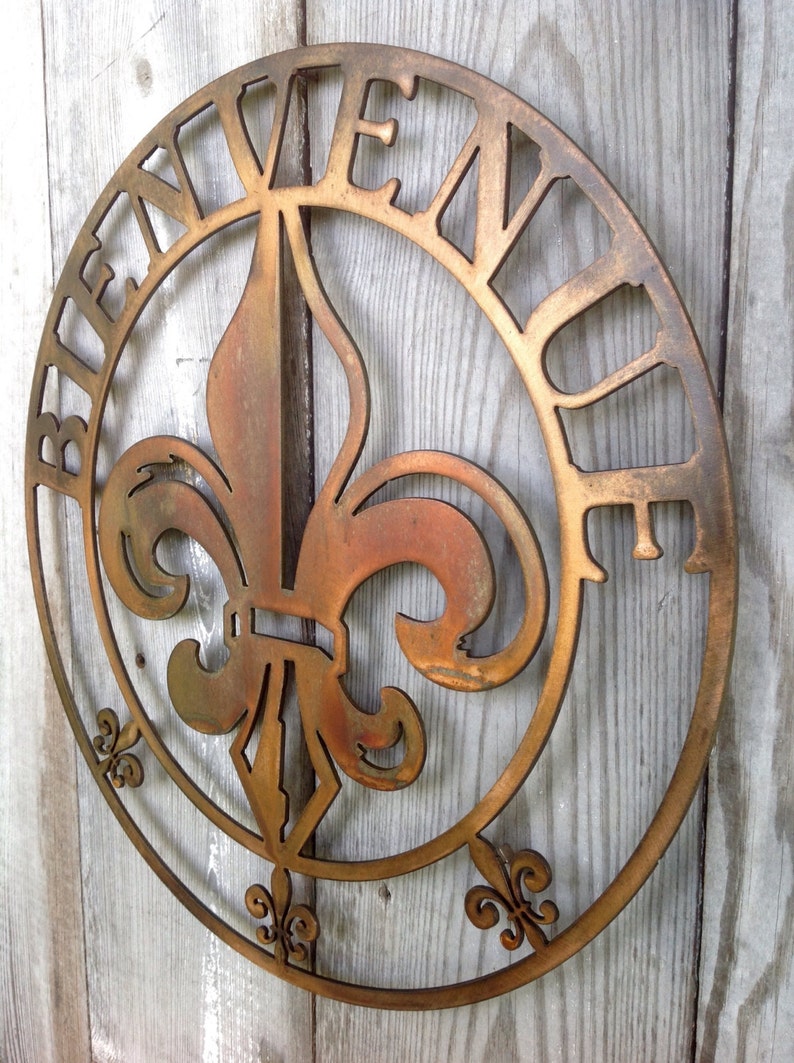 May include: A round metal sign featuring a fleur-de-lis design in the centre and the word "Bienvenue" around the edge. The sign is painted in a gold colour with a distressed finish.
