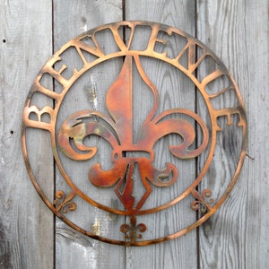 May include: A copper-coloured metal sign with a fleur-de-lis design in the centre and the word "Bienvenue" around the edge.