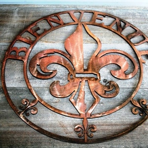 May include: A round metal sign featuring a fleur-de-lis design and the word "Bienvenue" inscribed around the edge. The sign is a rusty copper colour and has a distressed appearance.