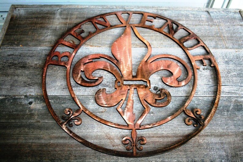 May include: A round metal sign featuring a fleur-de-lis design and the word "Bienvenue" inscribed around the edge. The sign is a rusty copper colour and has a distressed appearance.