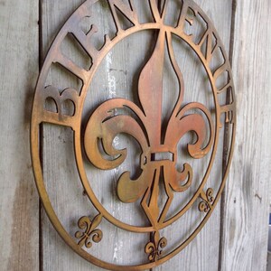 May include: A round metal sign featuring a fleur-de-lis design in the centre. The sign is painted in a rusty brown colour and has the word "Bienvenue" written around the edge.