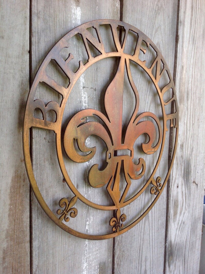 May include: A round metal sign featuring a fleur-de-lis design in the centre. The sign is painted in a rusty brown colour and has the word "Bienvenue" written around the edge.