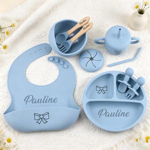 May include: A light blue silicone feeding set featuring a bib, a divided plate, a bowl, a lidded cup with a straw, and utensils. The bib, plate, and wooden-handled spoon and fork are personalized with the name Pauline and a bow graphic.