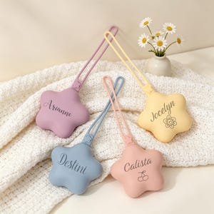 May include: Four star-shaped silicone keychains with loops, each featuring a unique name: Arianne in purple, Destini in blue, Calista in pink with a cherry icon, and Jocelyn in yellow with a smiley flower icon, all arranged together.