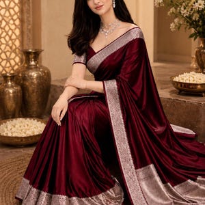 May include: A deep burgundy satin saree featuring a wide, ornate silver metallic border with intricate floral patterns. The garment includes a matching short-sleeved blouse with a silver border along the sleeves and a V-neckline, draped elegantly.