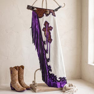 Pode incluir: Chaps de couro branco com longas franjas roxas, detalhes em couro castanho com gravação floral e uma aplicação de cruz. O cós inclui um concho de caveira e o texto ROCK. Um par de botas de cowboy castanhas com saltos roxos está próximo.