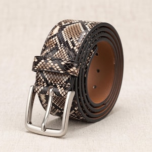 May include: A coiled belt featuring a black and beige snakeskin pattern on the exterior surface. The belt includes a silver-toned metal rectangular buckle and a matching loop. The interior side of the belt is a solid, smooth brown color.