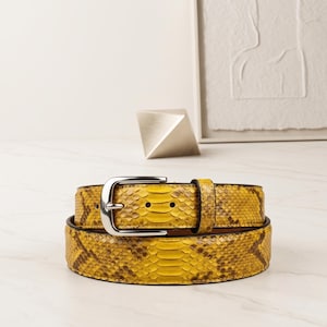 May include: A yellow and brown snakeskin patterned belt featuring a silver-toned metal buckle. The belt has a textured scale finish and a matching leather keeper loop, with a standard prong closure system and visible adjustment holes.