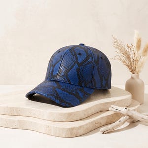 May include: A baseball cap featuring a vibrant blue and black snakeskin pattern. The cap has a structured crown, a curved brim, and a small button at the top centre. The material displays a textured, reptilian scale design throughout the exterior.