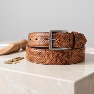 Puede incluir: Un cinturón marrón con textura de piel de serpiente y una hebilla de metal en tono plateado mate. El cinturón presenta un patrón de escamas repetitivo en tonos bronceado y marrón oscuro, con una trabilla a juego y cierre de hebilla.
