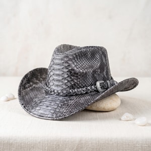 May include: A grey cowboy hat featuring a detailed snakeskin texture across the entire surface. The hat includes a braided band around the crown, accented with a metallic buckle detail. The brim is curved upward on the sides in a classic style.