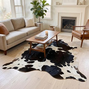 Könnte beinhalten: Ein großer, unregelmäßig geformter Kuhfellteppich mit schwarz-weißem Fleckenmuster. Der Teppich liegt flach auf einem hellen Holzboden unter einem rechteckigen Couchtisch aus Holz, der über eine Ablage und mehrere gestapelte Bücher verfügt.