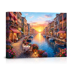 May include: A canvas print depicts a picturesque canal scene in Venice at sunset. Buildings line the waterway, adorned with flowers and awnings. Boats float on the water, reflecting the warm hues of the sky. A table and chairs are set up on the cobblestone path.