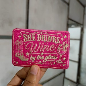 Può includere: Una toppa rettangolare ricamata in rosa acceso con cuciture dorate che riporta la scritta "SHE DRINKS Wine by the glass". Il design include uno stivale da cowboy ricamato a sinistra, un calice di vino a destra e piccole stelle decorative.