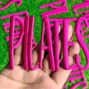 May include: A hot pink embroidered "PILATES" patch held in a hand, with other similar patches scattered on a green surface. The letters are in a cursive font, and the embroidery has a textured appearance.