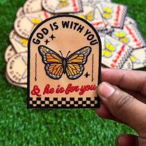 Könnte beinhalten: Gestickter Aufnäher mit einem Schmetterlingsdesign und dem Text "GOD IS WITH YOU & he is for you." Der Aufnäher hat einen beigefarbenen Hintergrund, einen schwarzen Rand und ein Schachbrettmuster am unteren Rand. Der Schmetterling hat orange und schwarze Flügel.