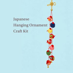 May include: A Japanese hanging ornament craft kit featuring a vertical string of small, multi-colored fabric shapes in red, blue, yellow, and green. The ornament is topped with a gold decorative knot and ends with a small gold bell at the bottom.