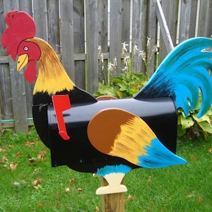 May include: A handcrafted mailbox shaped like a rooster. The mailbox is painted black with a red comb, yellow and brown neck, and blue tail feathers. A red flag indicates mail is present. The mailbox is mounted on a wooden post in a grassy yard.