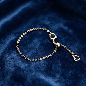 May include: A gold-toned chain bracelet featuring a circular spring ring clasp and an adjustable sliding bead mechanism. A small, open-outline heart charm hangs from the end of the chain, which extends beyond the sliding bead closure.