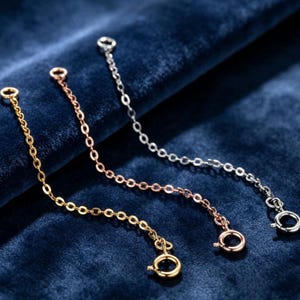 May include: Three jewelry extender chains in gold, rose gold, and silver tones. Each chain features a series of small oval links and terminates with a circular spring ring clasp at one end and a small jump ring at the opposite end for attachment.