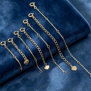 May include: A set of five gold-toned jewelry extender chains of varying lengths, each featuring a circular spring ring clasp at one end and a small, flat heart-shaped charm at the other end, arranged in a diagonal line from shortest to longest.