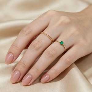 Puede incluir: Dos anillos de oro en los dedos. Uno presenta un delicado diseño de eslabones de cadena, mientras que el otro es una banda de oro fina y lisa rematada con una pequeña piedra preciosa redonda de color verde vibrante en un engaste sencillo.