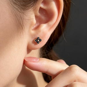 May include: A stud earring featuring four small, round, black stones arranged in a flower-like clover shape. The stones are set in a gold-toned metal frame with a delicate beaded edge, creating a compact and symmetrical design on the earlobe.