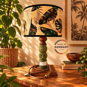 Peut inclure: Une lampe de table avec un abat-jour cylindrique orné d'un motif de feuilles tropicales vertes et brunes. La base est une figurine tiki en céramique verte avec un sourire aux dents blanches, surmontée d'un accent en bois brun.