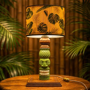 Könnte beinhalten: Dekorative Tischlampe mit Tiki-Stil-Sockel und gemustertem Lampenschirm. Der Sockel ist grün mit geschnitzten Gesichtsmerkmalen und gestapelten Elementen. Der Lampenschirm hat einen gelben Hintergrund mit schwarzen Blattmustern. Die Lampe steht auf einem Holztisch.