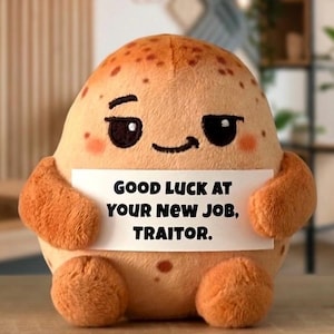 May include: A tan, round, plush potato toy with brown spots, embroidered black eyes, and a small smile. It holds a white rectangular sign with the black printed text: GOOD LUCK AT YOUR NEW JOB, TRAITOR. The plush has two small arms and two feet.