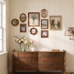 May include: A display of framed vintage art and photographs on a cream wall. Below, a dark wood dresser with six drawers is topped with vases of flowers. A floral lamp stands to the right.