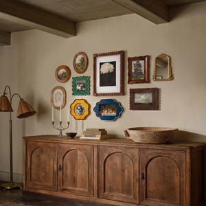 May include: A collection of framed art pieces and a mirror are displayed on a wall above a wooden cabinet. The frames vary in shape and color, with artwork depicting landscapes, still lifes, and portraits. A floor lamp with two shades stands to the left.