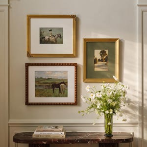 May include: Three framed art prints displayed on a wall above a marble console table. The top left print shows a person riding a horse, the bottom left shows two horses in a field, and the right print depicts a tree-lined path. A glass vase holds white flowers.