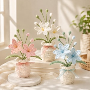 May include: Three crocheted flower arrangements in small cream-colored pots. Each pot contains two crocheted lilies in pink, white, or light blue, with green stems and buds. Each pot is tied with a thin ribbon matching the color of the flowers.