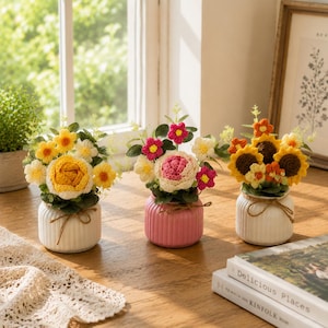 Puede incluir: Tres pequeños arreglos en maceta con flores de ganchillo en tonos amarillo, rosa y naranja. Cada arreglo se presenta en una maceta de cerámica acanalada, dos blancas y una rosa, decoradas con un fino lazo de cuerda marrón atado alrededor.