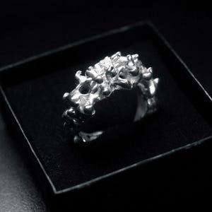 Puede incluir: Un anillo plateado con un diseño orgánico y texturizado, presentado en una caja negra. La superficie del anillo está cubierta de formas y protuberancias irregulares, creando una apariencia única y escultural. El anillo es probablemente de metal.