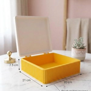 Puede incluir: Una caja de almacenamiento cuadrada de color amarillo con una tapa blanca con bisagras. El exterior presenta una textura acanalada vertical. La caja mide 7,5 cm de alto, 21 cm de profundidad y 20 cm de ancho, según las dimensiones indicadas.
