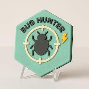 May include: A teal, hexagonal badge with the words "BUG HUNTER" in black lettering. The badge features a target design with a black bug silhouette in the center and a yellow lightning bolt. The badge is displayed on a clear stand.