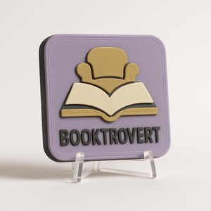 May include: A square lavender coaster featuring a raised graphic of a tan armchair positioned above an open book. The word BOOKTROVERT is embossed in black capital letters at the bottom. The coaster is displayed on a clear plastic stand.