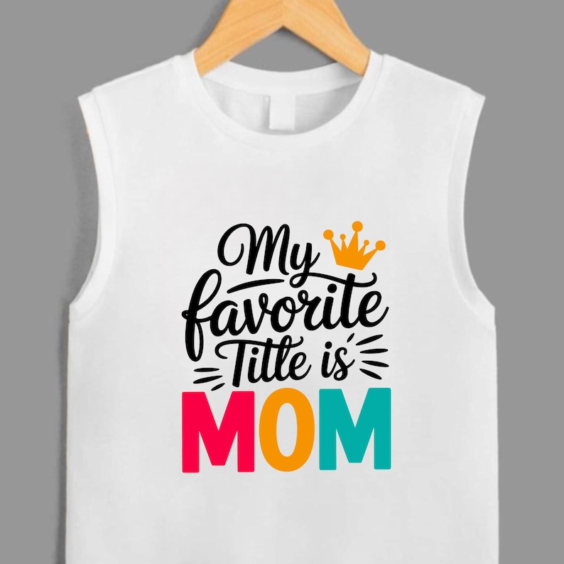 May include: A white sleeveless shirt featuring the black text "My favorite Title is" with an orange crown icon above it. Below, the word "MOM" is printed in large, bold letters with each character in a different color: pink, orange, and teal.